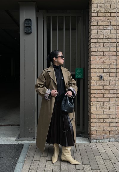Mujer que lleva abrigo beige tipo gabardina, falda negra plisada, botas beige, gafas de sol y sostiene un bolso negro, de pie junto a un muro de ladrillos y una puerta metalica.