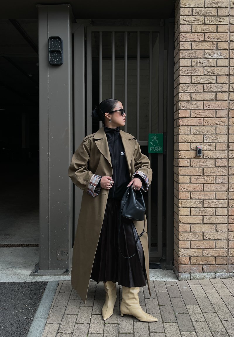 Frau trägt beige Trenchcoat, schwarzen plissierten Rock, beige Stiefel, Sonnenbrille und hält eine schwarze Handtasche, steht an einer Ziegelmauer und einem Metalltor.