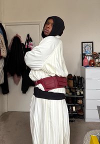 Personne vêtue d'une tenue blanche plissée et d'une capuche noire, debout de profil dans une pièce avec des manteaux, des chaussures et une commode avec des objets décoratifs derrière.