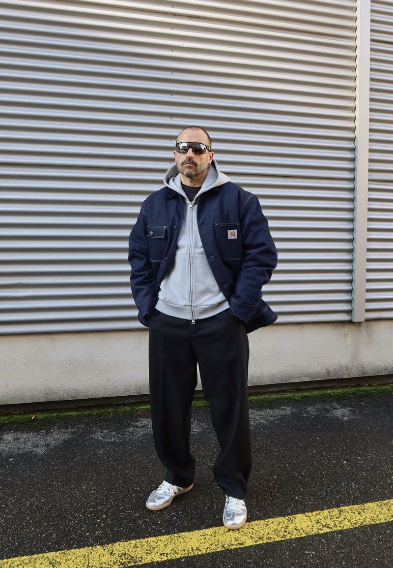 Homme portant des lunettes de soleil noires, une veste bleu marine, un sweat à capuche gris, un pantalon noir et des baskets blanches, debout sur l'asphalte avec un mur en métal ondulé en arrière-plan.