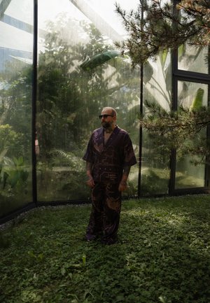 Conjunto de estilo kimono de color púrpura con un patrón y mangas cortas, de pie en un espacio verde exuberante rodeado de paredes de vidrio y plantas visibles.