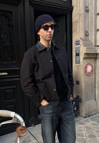 Άνδρας με μαύρο μπουφάν, μπλε σκουφί και γυαλιά ηλίου, με τα χέρια στις τσέπες, στέκεται κοντά σε μια μαύρη πόρτα και έναν πέτρινο τοίχο με θυροτηλέφωνο.