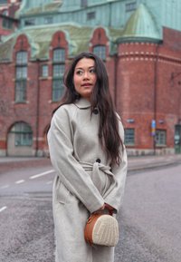 Light gray, wool blend coat with a belted waist and large black buttons. Rounded, woven handbag in tan and cream. Urban backdrop with brick building.
