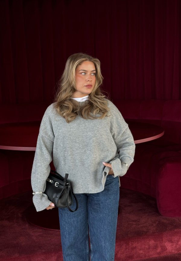 Ung kvinde med langt blondt hår iført en grå sweater og blå jeans, holder en sort håndtaske og står ved en mørkerød buet bænk.