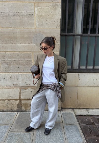 Mujer con blazer marrón oversize, camiseta blanca, pantalones deportivos grises, camisa de cuadros atada a la cintura, mocasines negros y gafas de sol, junto a una pared de piedra.