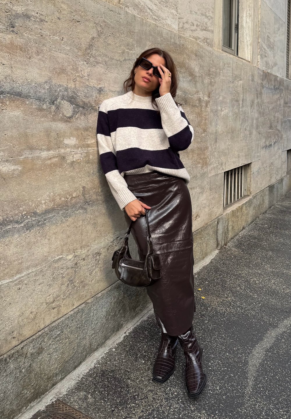 Donna che indossa un maglione a righe bianche e nere, una gonna in pelle marrone scuro e stivali, mentre tiene una piccola borsa nera, in piedi su una strada accanto a un muro.