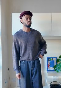 Kötött, sötétszürke pulóver bordás textúrával; kényelmes, laza szabás. Bő, kék farmernadrág. A bordó barett kiemeli az öltözéket semleges háttér előtt.