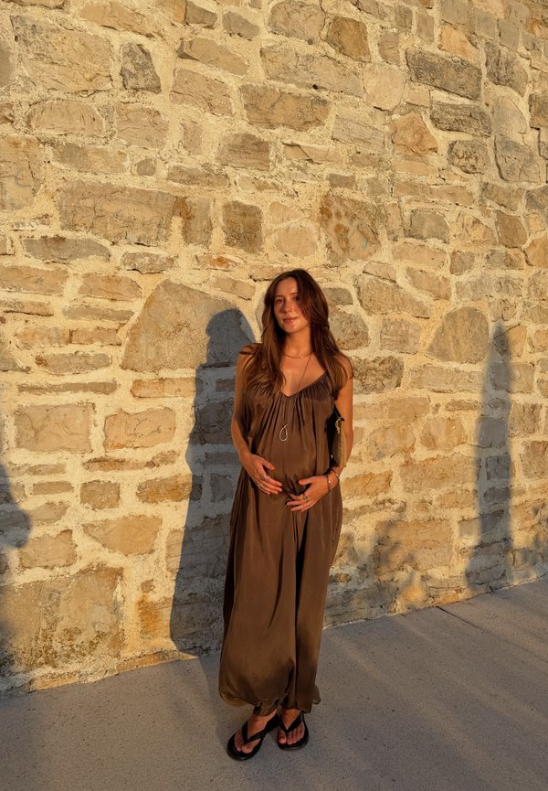 Femme en longue robe marron et sandales se tient contre un mur de pierre, tenant son ventre de femme enceinte, baignée de lumière solaire chaude.