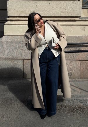 Mujer con abrigo largo beige, top blanco abotonado, vaqueros anchos oscuros, botas negras, gafas de sol, sosteniendo bolso blanco y teléfono cerca de una pared de piedra.