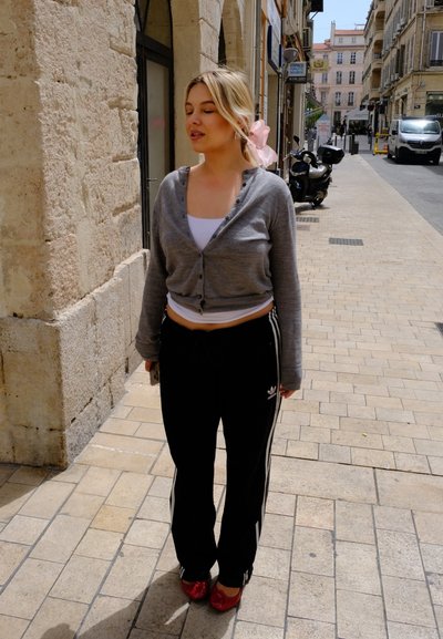 Cardigan gris, camiseta sin mangas blanca, pantalones de chándal negros con rayas blancas y zapatos rojos. Entorno al aire libre con paredes de piedra y callejón de adoquines.