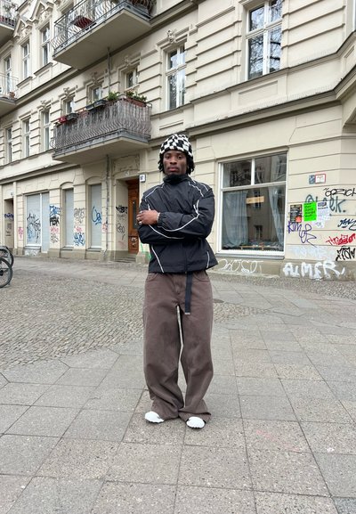 Rompevientos negro con rayas blancas, pantalones anchos marrones y un gorro de cuadros en blanco y negro en una calle de adoquines con grafitis.