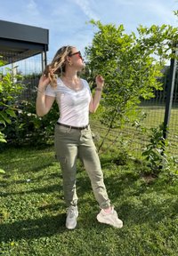 Fehér, kerek nyakú póló, olivazöld cargo nadrág oldalsó zsebekkel, és fehér sportcipő rózsaszín részletekkel, zöld háttér előtt.