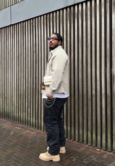 Camisa de cuadros beige, camiseta blanca, pantalones negros con detalle de cadena, botas color crema. De pie frente a un fondo de panel de madera.