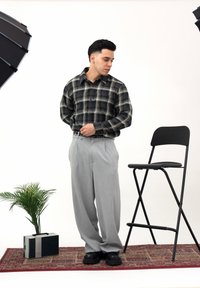 Chemise à carreaux noire et verte, pantalon plissé gris, ceinture noire et chaussures noires. Décor en studio avec une plante en pot et une chaise pliante.