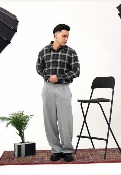Camisa de cuadros negra y verde, pantalones plisados grises, cinturón negro y zapatos negros. Escenario de estudio con una planta en maceta y una silla plegable.