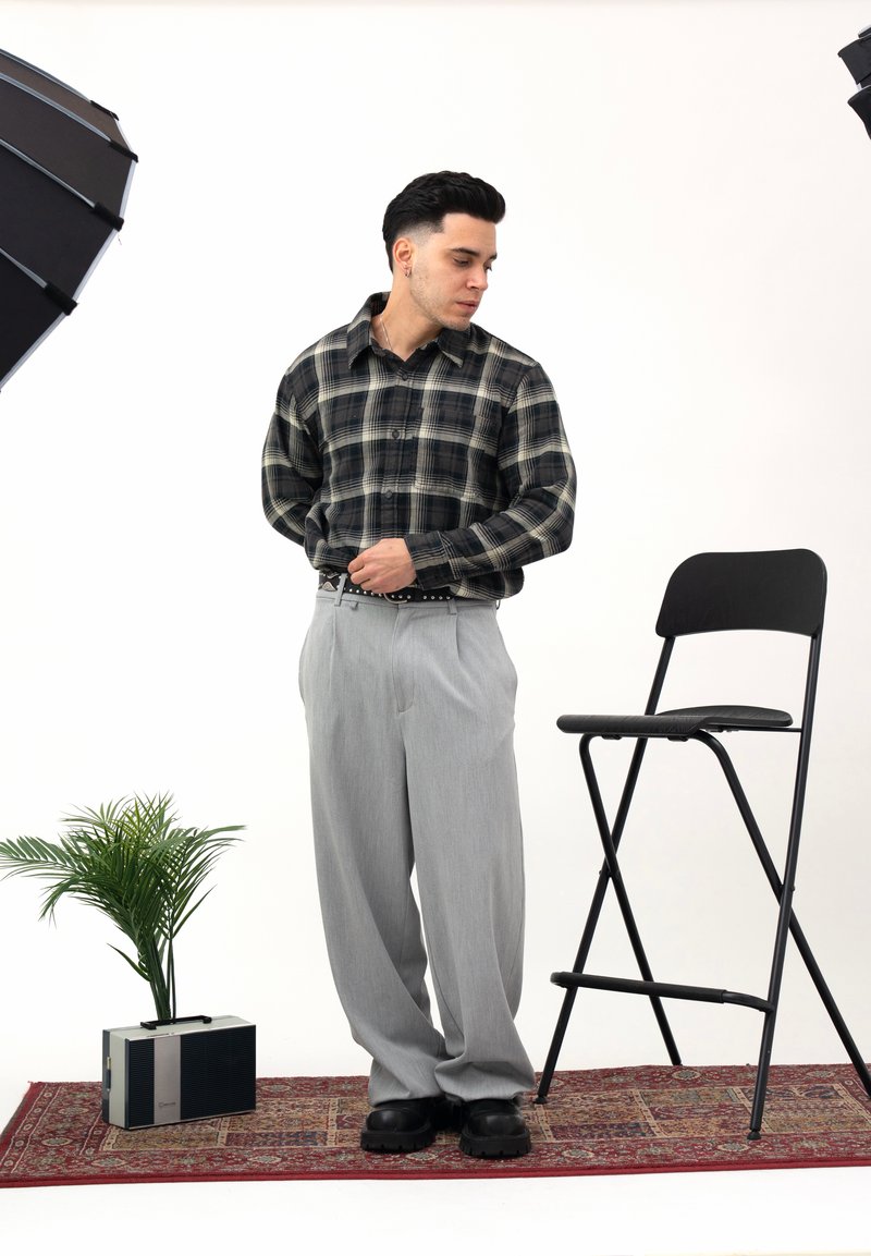 Schwarz-grünes kariertes Hemd, graue plissierte Hose, schwarzer Gürtel und schwarze Schuhe. Studio-Setting mit einer Topfpflanze und einem Klappstuhl.