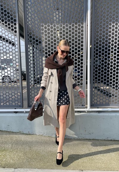 Mujer caminando al aire libre con un abrigo trench beige, suéter oscuro con bufanda, falda de lunares, zapatos negros, gafas de sol y llevando un bolso texturizado.