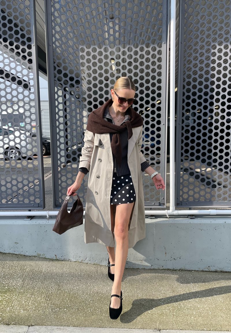 Mulher caminhando ao ar livre, vestindo um casaco trench coat bege, suéter escuro com lenço, saia de bolinhas, sapatos pretos, óculos de sol e carregando uma mala texturizada.