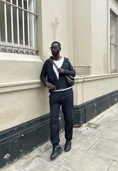 Chaqueta y pantalones deportivos negros, camiseta blanca debajo, zapatos negros con acentos rojos, de pie casualmente contra una pared.