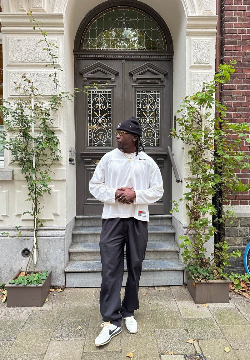 Λευκό πουκάμισο με μακριά μανίκια και στρογγυλή λαιμόκοψη, μαύρο παντελόνι και λευκά αθλητικά παπούτσια. Στεκόμενος μπροστά από μια ξύλινη πόρτα με διακοσμητικό γυαλί.