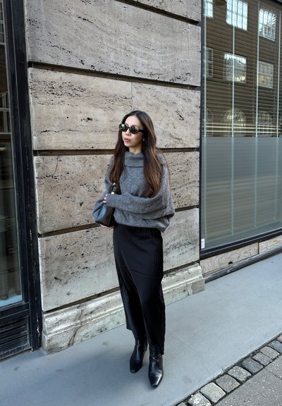 Suéter gris de gran tamaño, falda plisada negra y botas negras de tobillo. Los accesorios incluyen gafas de sol y un pequeño bolso negro. Fondo de pared de piedra.