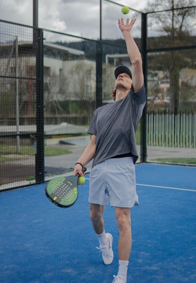 Raqueta de pádel con empuñadura verde y negra en la mano; una pelota de tenis amarilla está encima, en una pista azul. Sujetos con ropa casual en gris y azul claro.