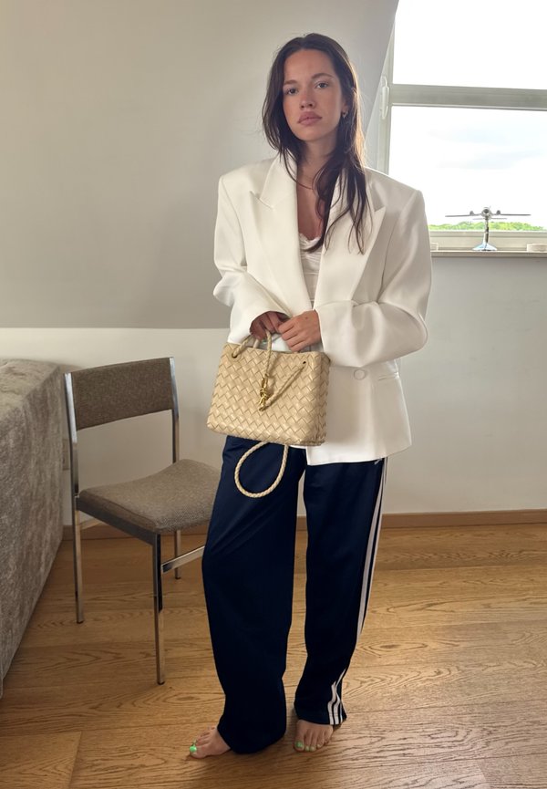 Bílý oversized blazer přes světlý top, námořnické jogger kalhoty s bočními pruhy a hnědá tkaná kabelka s krátkým uchem. Viditelná dřevěná podlaha.