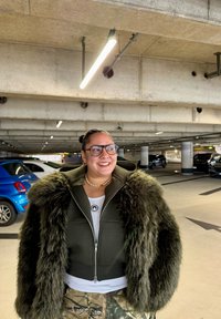 Veste en fausse fourrure verte avec fermeture éclair, superposée sur un haut blanc. En arrière-plan, un parking avec des voitures bleues et noires.