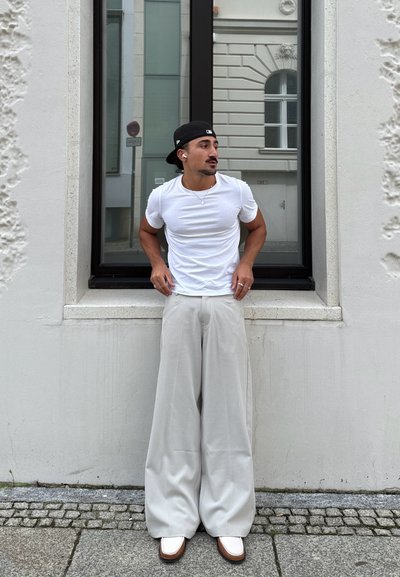 Camiseta blanca ajustada, pantalones palazzo sueltos de color gris claro y zapatos blancos con acentos marrones. El fondo presenta un edificio de color claro.