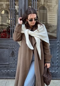 Femme en manteau marron et jean bleu clair ajuste ses cheveux, porte des lunettes de soleil et une écharpe blanche drapée sur les épaules, tenant un sac tissé marron.