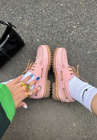 Rosa Lederschuhe mit kontrastierenden braunen Sohlen und Schnürsenkeln, ausgestattet mit Metallösen. Begleitet von einer schwarzen Tasche und weißen Socken mit Logo.