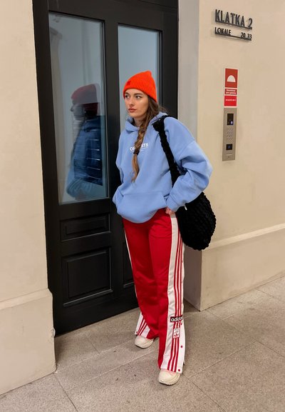 Sudadera azul clara con bolsillo frontal, joggers rojos con rayas blancas, bolsa negra de punto y gorro naranja. De pie cerca de una puerta negra.