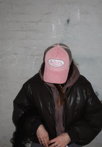 Hnedá kožená bomber bunda s ružovou kordovou čiapkou s bielym logom. Pod ňou šedá mikina, čierny textúrovaný doplnok a strieborné prstene.