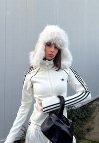 Γυναίκα που φοράει λευκό καπέλο με γούνα στο εσωτερικό, λευκό μπουφάν Adidas με μαύρες ρίγες, κρατώντας μια μαύρη τσάντα μπροστά σε γκρίζο τοίχο.