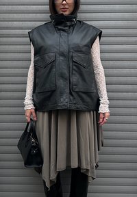 Czarna skórzana kamizelka bez rękawów z podniesionym kołnierzem i dwoma przednimi kieszeniami, noszona na koronkowej białej bluzce z długim rękawem oraz zwiewnej beżowej spódnicy.