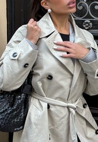Femme portant un trench-coat beige et des boucles d'oreilles en perles, ajustant le col de son manteau avec des ongles manucurés, portant un sac à main en cuir tressé noir.