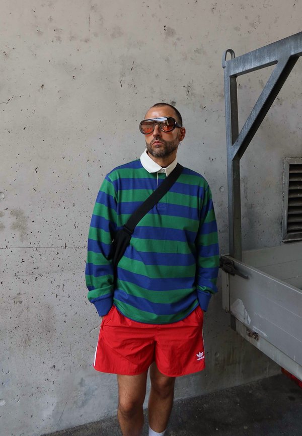 Maglietta a maniche lunghe a strisce verdi e blu, pantaloni corti rossi e occhiali da sole marroni oversize. Borse a tracolla nere e calzini bianchi con scarpe.