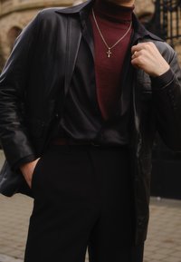 Zwarte leren jas over een donker shirt en een bordeauxrode coltrui, met een zilveren kruis ketting, zwarte broek en een bruine riem.