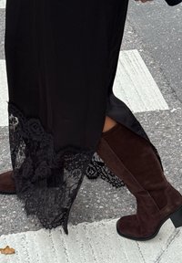 Schwarzes Kleid mit Spitzendetails am Saum, kombiniert mit knielangen Stiefeln aus dunkelbraunem Wildleder, die einen klobigen Absatz und eine strukturierte Oberfläche aufweisen.