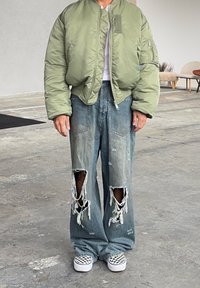 Olivově zelená bomber bunda, volné světlé modré roztrhané džíny s velkými dírami a černo-bílé boty na nazouvání s károvaným vzorem.