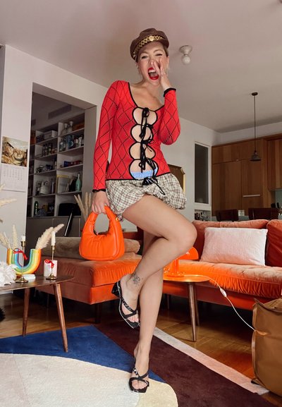 Bodysuit rojo de manga larga con patrón de diamantes y detalles de encaje negro; falda de cuadros; bolso naranja; sandalias negras; entorno interior con decoración colorida.