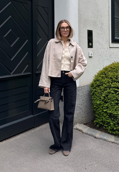Chaqueta beige corta sobre una blusa de punto crema, combinada con jeans negros acampanados y zapatos grises; sosteniendo un pequeño bolso gris.