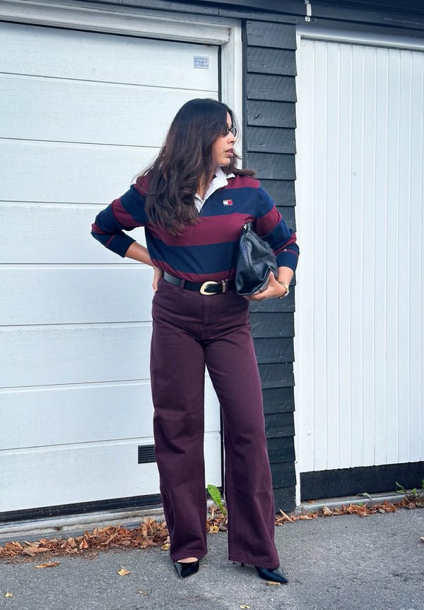 Femme portant un haut rayé bordeaux et bleu marine et un pantalon bordeaux, tenant un sac noir, debout devant des portes de garage blanches regardant sur le côté.