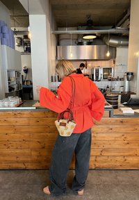 Φωτεινό πορτοκαλί χαλαρό τζάκετ, σκούρα φαρδιά τζιν και μια μικρή τσάντα με σχέδιο. Ξύλινο πάγκ ο και φωτιστικά σε καφετέρια.