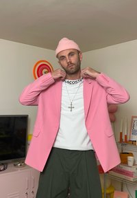 Ljusrosa blazer av lättviktsmaterial, bärs över en vit skjorta med en logotyp, matchas med lösa gröna byxor och en rosa mössa.