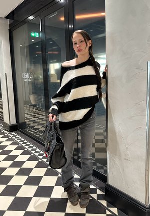 Suéter de rayas en blanco y negro de gran tamaño, jeans grises y calzado marrón. Sosteniendo un bolso gris. Diseño moderno y texturizado en un entorno interior.