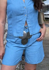Svetlo modra denim brezrokavnik s gumbi, v kombinaciji z ujemajočimi se hlačami. Siva dekorativna sponka za pas z zapletenim vzorcem.