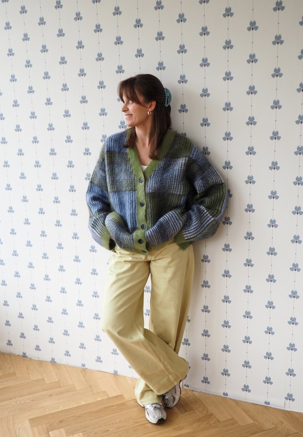 Mujer apoyada en una pared con papel pintado floral, lleva un cárdigan azul verdoso de cuadros, pantalones amarillos, zapatillas y una pinza para el pelo, sonriendo hacia un lado.