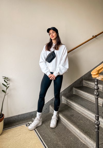 Una persona vestida con un sudadera blanca oversized, leggings negros, zapatillas blancas y un bolso cruzado negro está de pie en una escalera.
