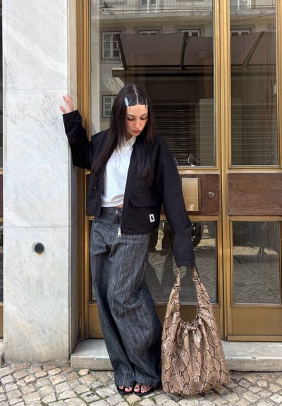 Mujer joven con cabello largo y oscuro, lleva chaqueta negra, camisa blanca, pantalones anchos grises, sostiene un bolso grande con estampado de serpiente, de pie junto a una puerta de cristal con marco de latón.